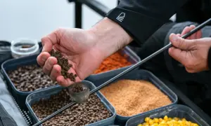 Ręka z pelletem, przygotowująca zanętę na ryby. W tle pojemniki z różnymi składnikami.