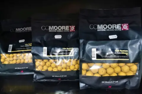 Trzy opakowania kulek proteinowych CC Moore Live System w różnych rozmiarach. Kulki są żółte, widoczne przez przezroczyste okienko.