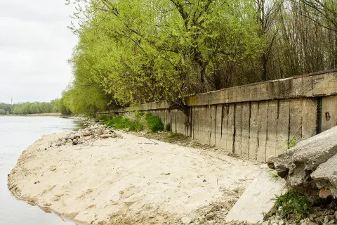 Betonowa opaska na rzece chroni brzeg przed erozją. Wiosenne drzewa tworzą zieloną ścianę nad wodą.