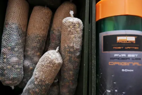Kilka woreczków z zanętą w siatce PVA, obok zielony pojemnik z pomarańczową nakrętką, na którym widnieje napis "PVA SET SPIDER WEB ORIGINAL".