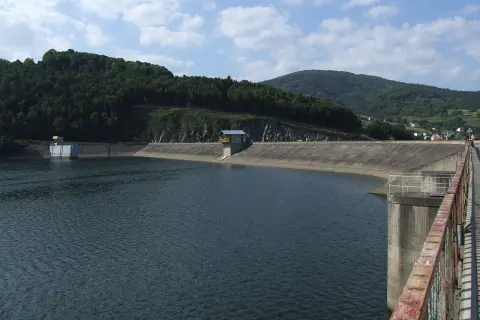 Betonowa zapora tworzy rozległe, spokojne tresna jezioro. W oddali widać zalesione wzgórza i niewielką wieś.