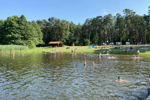 Słoneczny dzień nad jeziorem Szczutowo. Ludzie kąpią się, opalają na trawie i odpoczywają pod wiatą.