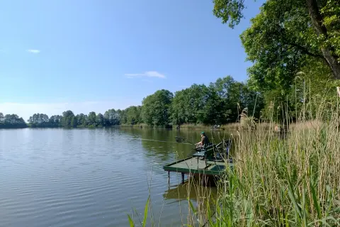 Wędkarz na pomoście nad spokojnym zalewem w Nowym Mieście. Otoczenie zieleni i błękitne niebo tworzą sielski krajobraz.