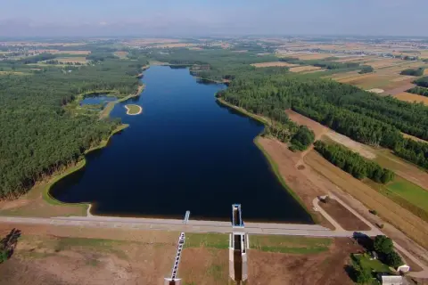 Widok z lotu ptaka na zalew Jagodno otoczony lasem i polami. Woda lśni w słońcu, a tama z budowlami hydrotechnicznymi stanowi imponującą konstrukcję.