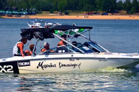 Łódź motorowa z pasażerami płynie po wodzie, tworząc pianę. Na burcie widnieje napis "Marina Domaniewska".