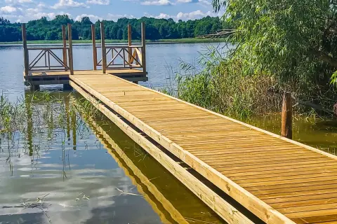 Drewniany pomost nad jeziorem zaprasza do spaceru. Woda odbija błękitne niebo i zielone drzewa.