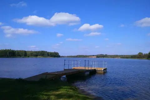 Drewniana przystań na spokojnym zalewie Drzewica. Błękitne niebo z białymi chmurami nad taflą wody i zielonymi lasami w tle.
