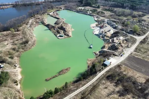 Widok z lotu ptaka na turkusowe jezioro zajączkowskie, otoczone piaskowymi skarpami i zielenią. Widać sprzęt wydobywczy i łódź.