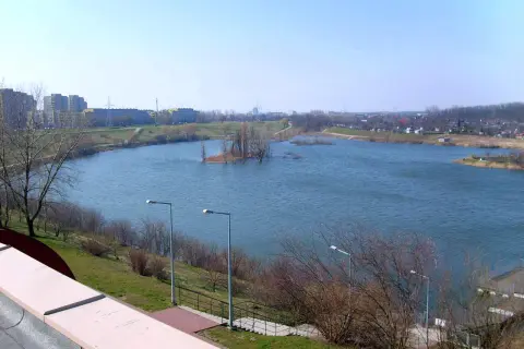 Spokojne jezioro Zatorze otoczone zielenią i zabudowaniami miejskimi. Na wodzie mała wyspa z drzewami.