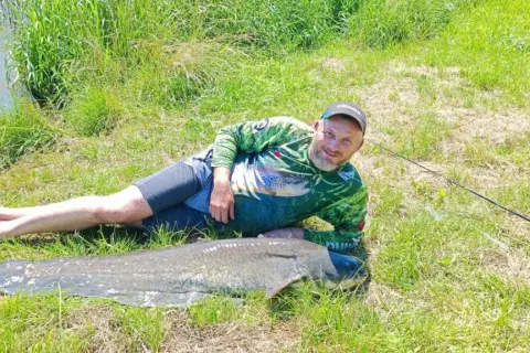 Szczęśliwy wędkarz z potężnym sumem złowionym w zbiorniku Nielisz. Obok łódka i wędka.
