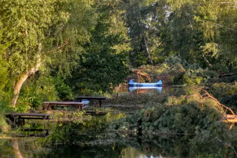 Niebieska łódź czeka na brzegu zwalonego drzewa. Jezioro zmarłe, otoczone bujną zielenią, odbija światło.