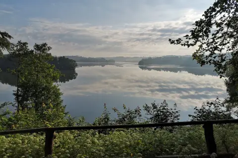 Spokojny poranek nad jeziorem w Sulęczynie. Lustrzana tafla wody odbija chmury i zalesione brzegi.