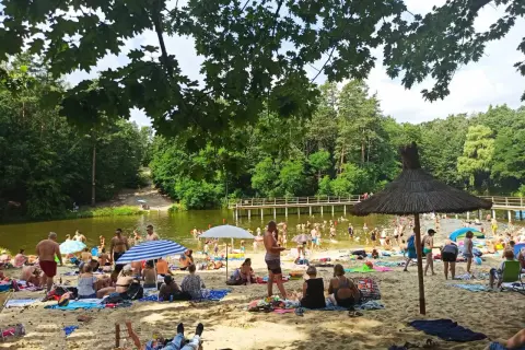 Słoneczny dzień na plaży przy zalewie w Garbatce. Ludzie odpoczywają na piasku, kąpią się w wodzie i spacerują po pomoście.