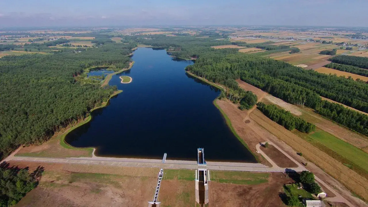 Widok z lotu ptaka na zalew Jagodno otoczony lasem i polami. Woda lśni w słońcu, a tama z budowlami hydrotechnicznymi stanowi imponującą konstrukcję.