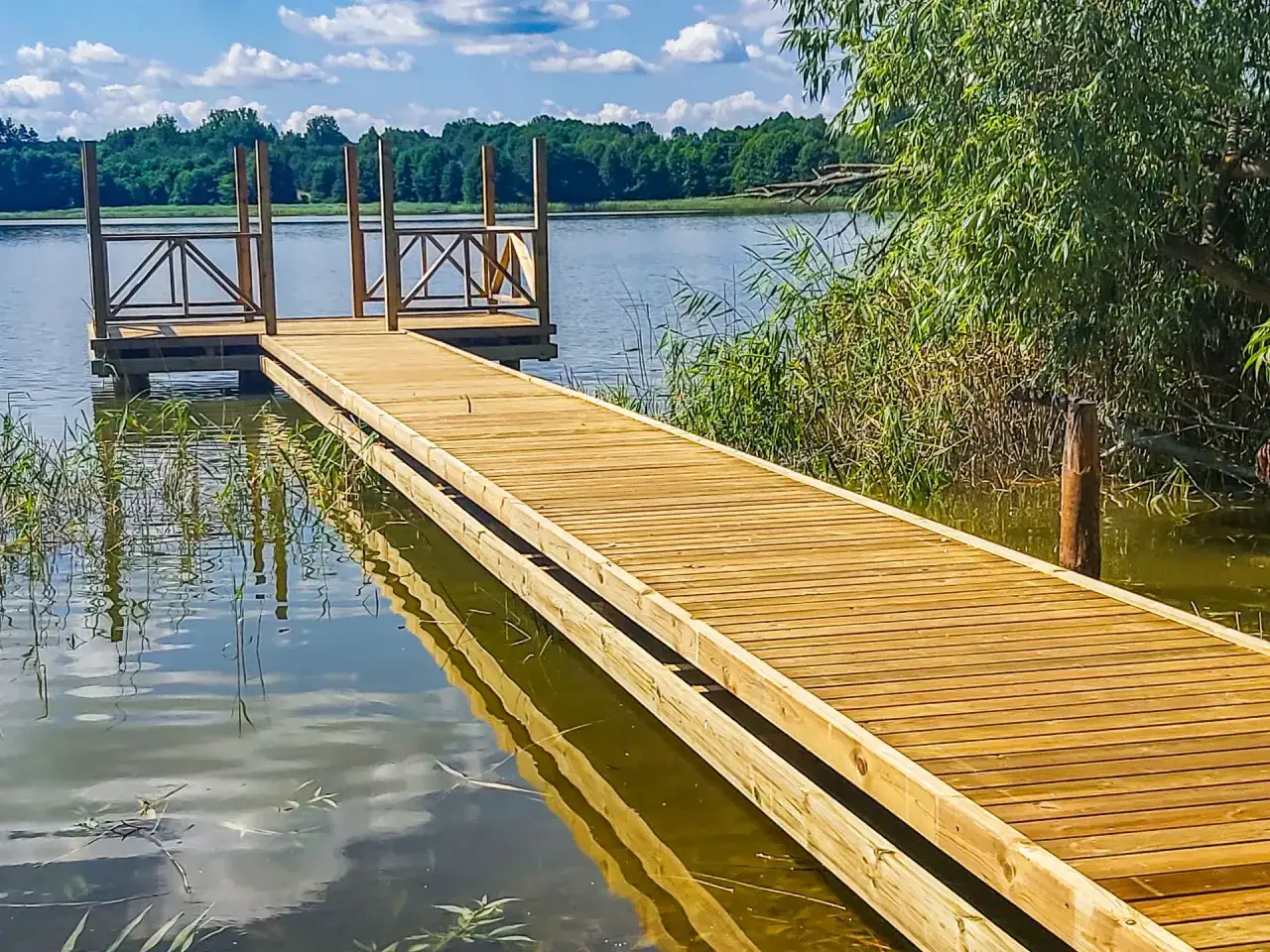 Drewniany pomost nad jeziorem zaprasza do spaceru. Woda odbija błękitne niebo i zielone drzewa.