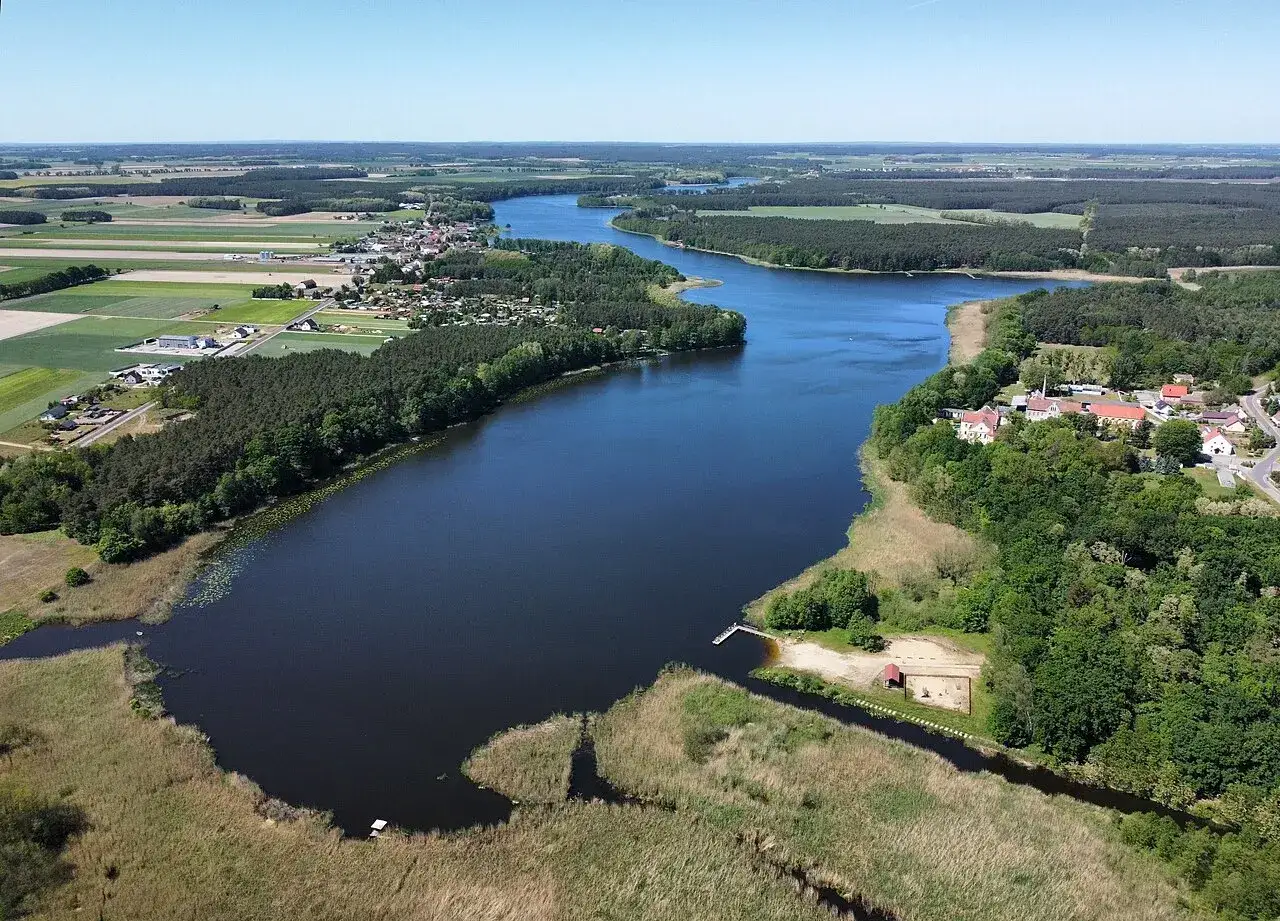 Malownicze jezioro wojnowskie otoczone lasami i polami. W oddali widać zabudowania wsi, a na brzegu plażę z placem zabaw.