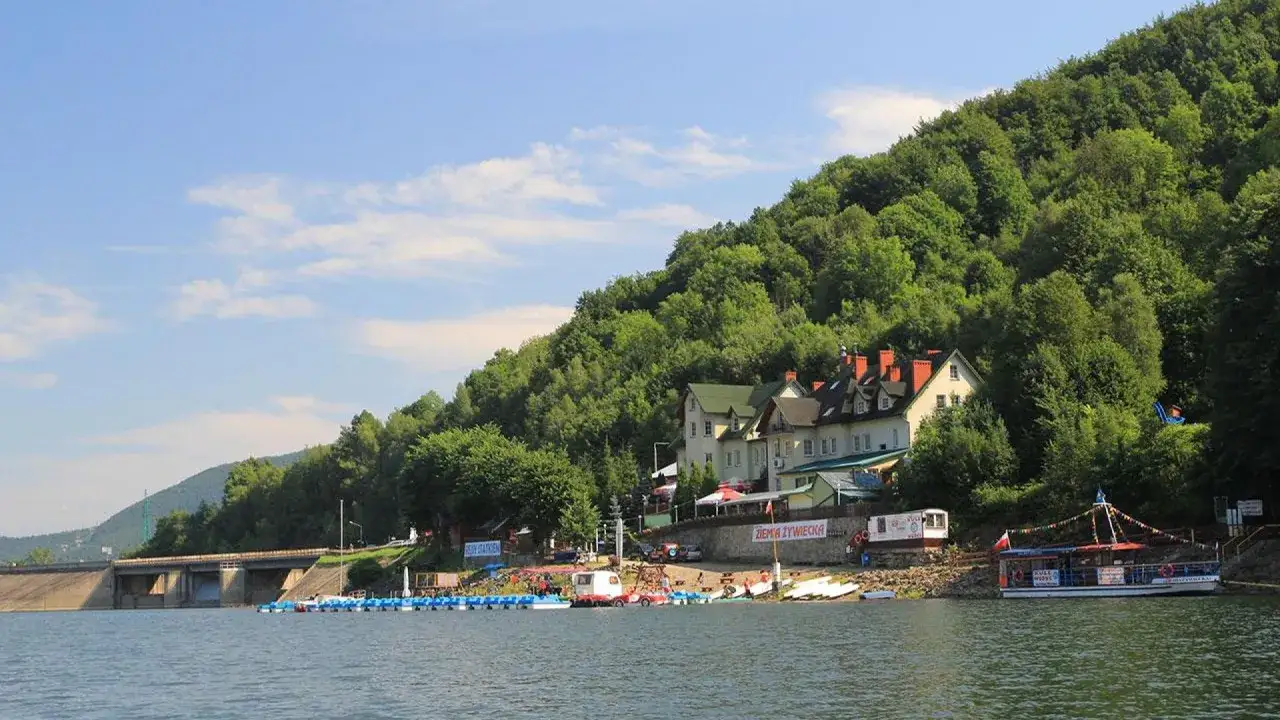 Malownicze jezioro Tresna otoczone zielonymi wzgórzami, z budynkami nad brzegiem i łodziami na wodzie.