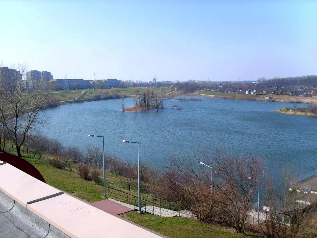 Rozległe jezioro Zatorze otoczone zielenią i zabudowaniami, z wyspą porośniętą drzewami pośrodku.