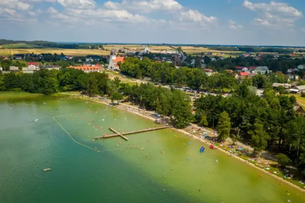 Tłum ludzi wypoczywa nad jeziorem wielewskim. Woda jest zielona, a na brzegu widać plażę i las. W oddali majaczy kościół.