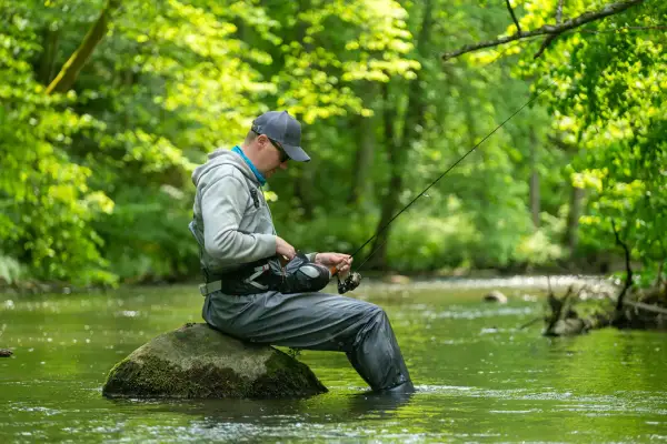 Wędkarz siedzi na kamieniu w rzece, przygotowując sprzęt. Zielone drzewa tworzą tło dla tej spokojnej przykosa na rzece.